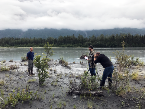 Three men are shown, one is filming, one is standing with a paper presumably asking questions, and the third is standing in the distance facing the camera. In the background of the photo is a river with trees and grey clouds above. 