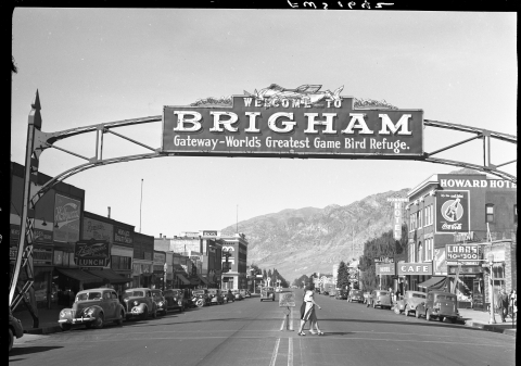 Large sign over a busy street in Brigham, Utah; the sign is supported by a truss-like metal structure, with support legs on either side of the street. The sign reads "Welcome to Brigham - Gateway - World's Greatest Game Bird Refuge.". The sign also features an illustration of waterfowl at the top of the sign. Numerous cars of 1940s vintage are parked along the street. Two people are passing directly underneath the sign, via the crosswalk. Mountains loom in the background.