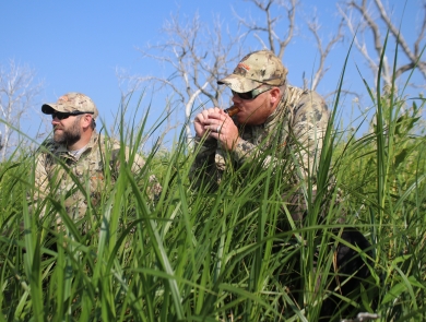 Two men dressed in camouflage sitting in grass. One man is holding a duck call to has mouth.