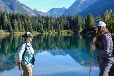 Two women wearing baseball caps and backpacks hold walking sticks while talking in front of a clear, still waterbody that is reflecting mountains and trees