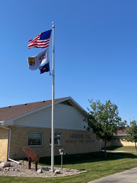 3 flags on flagpole with US flag atop USFWS ones. Building behind flagpole reads Garrison Dam National Fish Hatchery