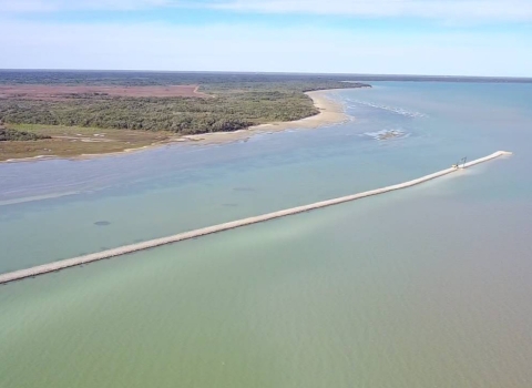 Aerial photo showing breakwater in progress along the coast. The breakwater is long and thin in the middle of blur water and the coast is green with some sandy beach.