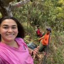 A woman on a steep wooded slope poses for a selfie. Two people smile in the background.