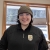 A woman in a green winter hat and a black jacket with the U.S. Fish and Wildlife Service logo on the chest. She wears large square glasses and stands in front of a window. 