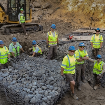 Ten people wearing bring yellow shirts and blue hard hats pose near a newly constructed rock wall