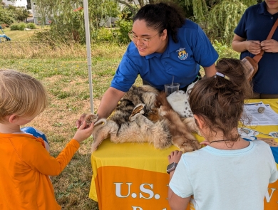 a woman in a blue USFWs uniform smiles while teaching kids about wildlife at an informational table outside