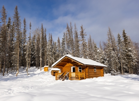 A wood cabin stands in the snow at the edge of a forest.