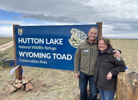 Two people posing for a photo outside in front of a sign that reads, “Hutton Lake National Wildlife Refuge Wyoming Toad Conservation Area”. The sign is dark blue and has the U.S. Fish and Wildlife Service logo on it and a picture of the Wyoming Toad