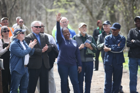 A smiling woman raises her arm high while a group of people standing around her smile at her and applaud.