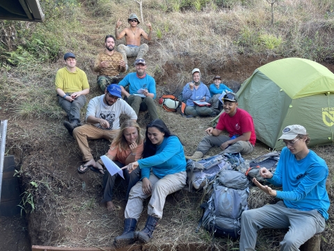 A group of young people sit along a grassy slope. Backpacks and a tent are nearby, and in the center a young woman points to a document her coworker is holding, while her other coworkers around her pantomime the motion or smile at the camera.. 