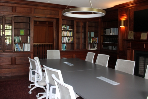 A modern conference style table sits in the center of a room with bookshelves and dark wood panels on the walls. 