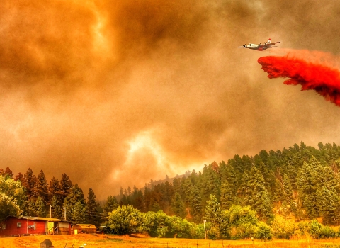 airplane drops red smoke in smoke-covered sky over trees, house