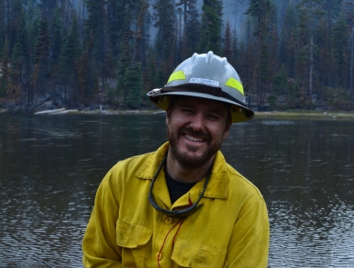 A firefighter dressed in yellow protective clothing and hard hat, smiles and leans on their tool with a radio in hand. Behind him is a mountain lake lined with tall evergreen trees. The sky is hazy with smoke.