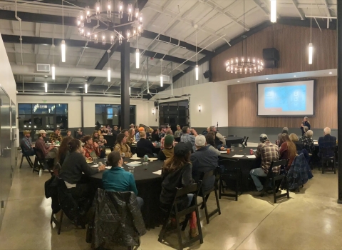 People sitting around round tables at a meeting with a speaker on a stage with a presentation up on a larger screen.