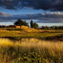 Sunset with clouds in the sky, building on top of hill overlooking wetland filled with grass and other plants 
