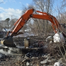 The orange arm of a construction excavator reaches into a river to break apart a dam