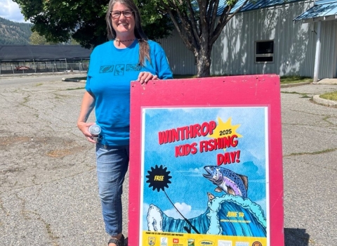A woman in a blue shirt stands next to a red sign reading "Winthrop Kids Fishing Day"