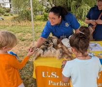 a woman in a blue USFWs uniform smiles while teaching kids about wildlife at an informational table outside