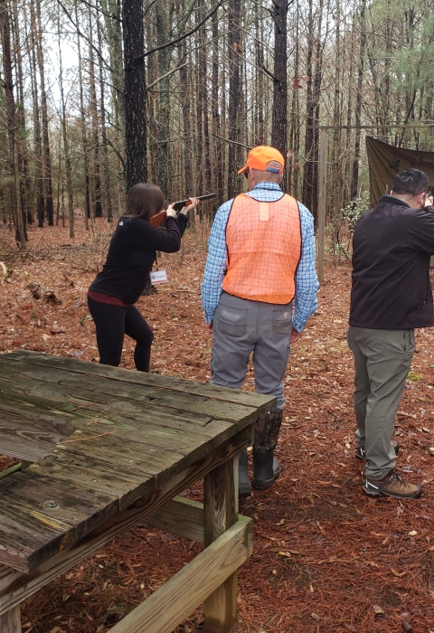  A man and a women holding firearms face the woods with a man with a bright orange vest on in between them.