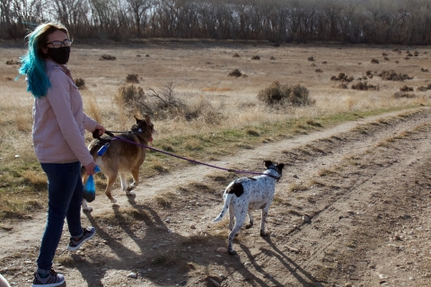 Woman walking down trail with two leashed. Sagebrush uplands, trees, and a lake can be seen in the distance.