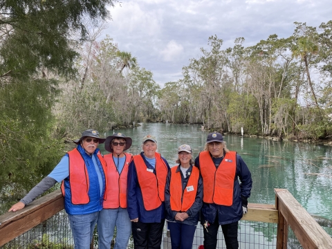 Five boardwalk volunteers in uniform standing on boardwalk overlook with manatees in the springs behind them. 