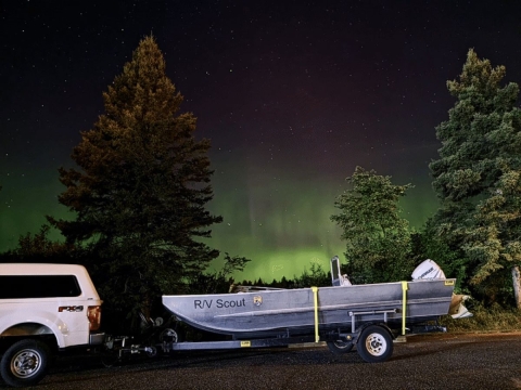 A truck pulling a boat is parked on the side of the road with the northern lights displaying in the sky