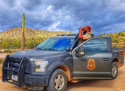 A law enforcement officer standing in his truck in a desert setting looking through binoculars