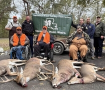 Photo of five harvested deer in the foreground and three hunters in wheelchairs in the middle and several individuals standing behind them with a green banner. 