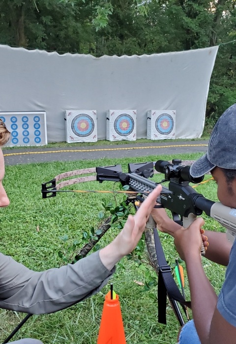 A man teaching a woman how to handle a crossbow while she aims at an archery target
