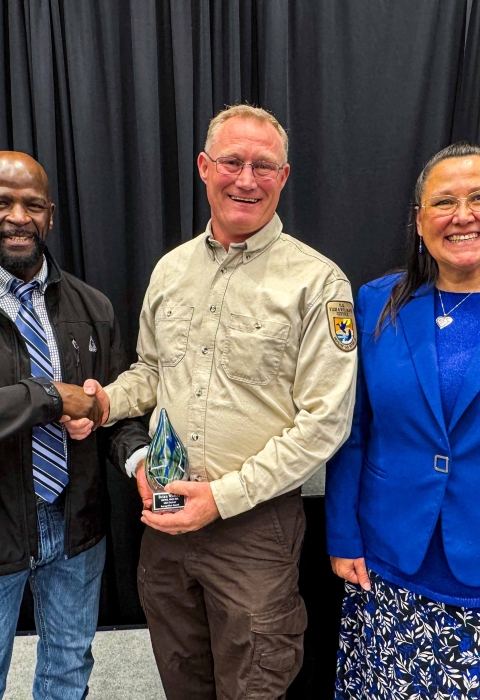 A unformed FWS employee shakes hands with another individual while holding an award. A woman stands smiling to the right of the awardee.