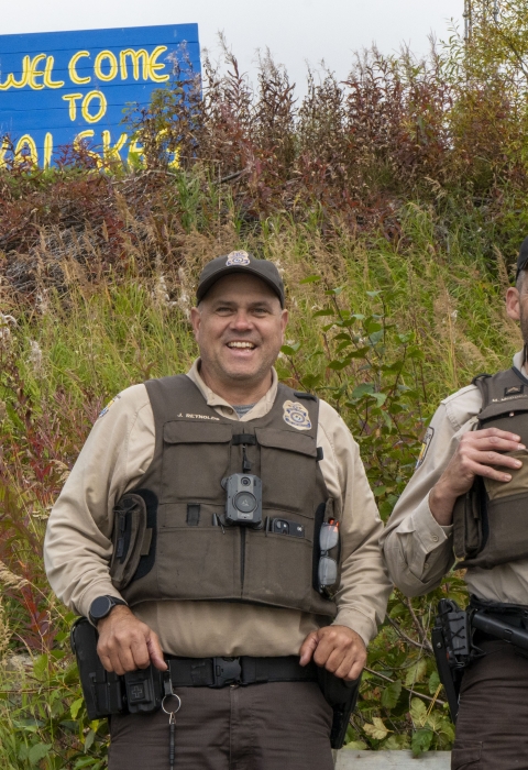 Two uniformed Federal Wildlife Officers pose for a picture with a painted sign in the background on a hill reading "welcome to Kalskag" 