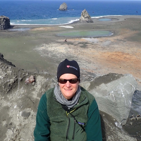 Woman in green shirt wearing stocking cap and sunglasses with brown island of and blue ocean behind her.