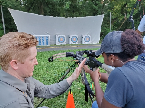 A man helps a woman handle a crossbow while she aims at an archery target.