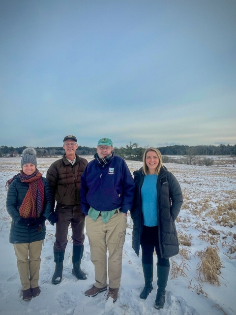 Four people stand in the foreground, with snowy land, cloudy sky, and a large green tree behind them