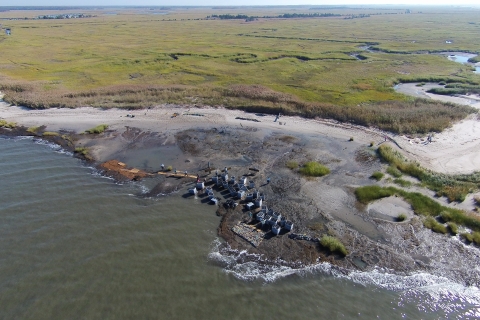 In this aerial image, Gandys Beach is visible in the foreground, as are a handful of volunteers and project staff and large square, gray containers filled with reef materials. A green marsh is visible behind the beach.