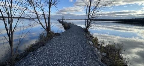 a stone jetty with sparse trees cuts through a still lake that reflects blue skies and clouds