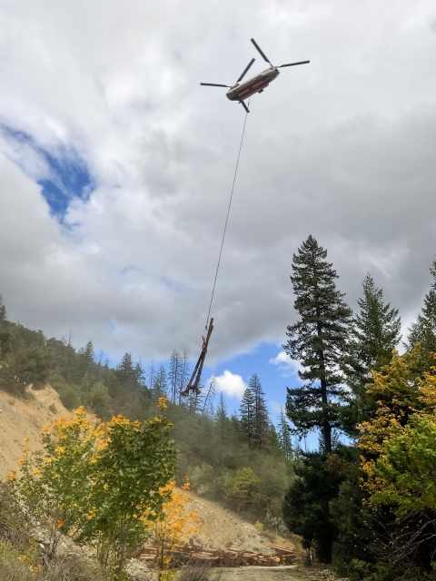 A chinook helicopter flies with a wood log attached to a rope. Mountains and trees are in the background. 