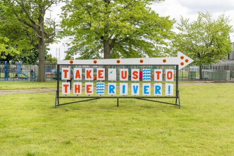 A large sign on green grass reads "Take us to the river"