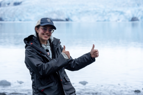 A young woman in a dark rain jacket smiles and holds up two thumbs up. She stands in front of icy water, with a large glacier visible in the background.