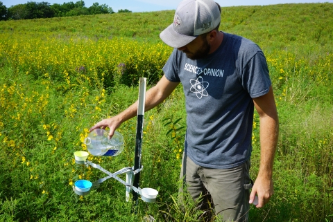 An individuals pours water into a man-made structure with 3 small bowls in a prairie