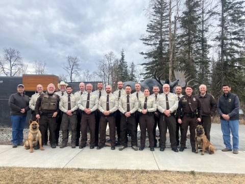 19 people pose for a picture in front of a statue of a bull moose. 17 of them are wearing Federal Wildlife Officer uniforms. There is an officer with a k9 on either side of the group.; 