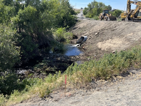 A small stream runs through a wooded area beside a dirt road. Construction equipment, including a backhoe, is parked near the road. Vegetation surrounds the stream, and a pipe juts out, discharging water into the stream.