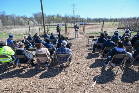 A group of people seated in an audience listening to a speaker with a fence and field in the background.