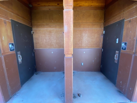 Image of two brown doors across from each other with a light brown divider wall between them. Men's and women's bathroom signs with the accessible symbol are next to each door.