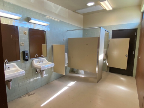 Image of a restroom interior with two white sinks and several feet of space in front of them. There are two mirrors over the sinks, a urinal behind a small tan divider, and a toilet behind tan walls and a large door.