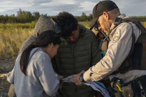 A uniformed Federal Wildlife Officer on a bank of a river showing a map to four moose hunters