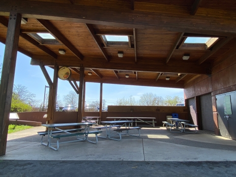 Image of several metal picnic tables on a concrete pad under a wooden roof with skylights.