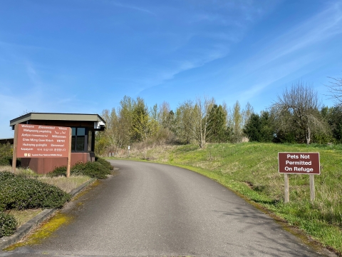 Image of an asphalt driveway passing by a sign that read pets not permitted and another sign reading welcome in many different languages. The driveway goes past a small brown booth structure with grass and trees surrounding it.