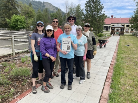 A group of 7 people stand in front of the pollinator garden holding a heritage garden sign