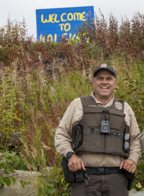 a uniformed officer poses in front of a sign that reads welcome to Kalskag